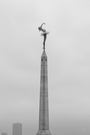 Black and white fine art ai photograph of a ballerina in a dramatic arched back pose on the tip of a tall spire, viewed from below, with high contrast and ethereal lighting accentuating her silhouette against the sky.