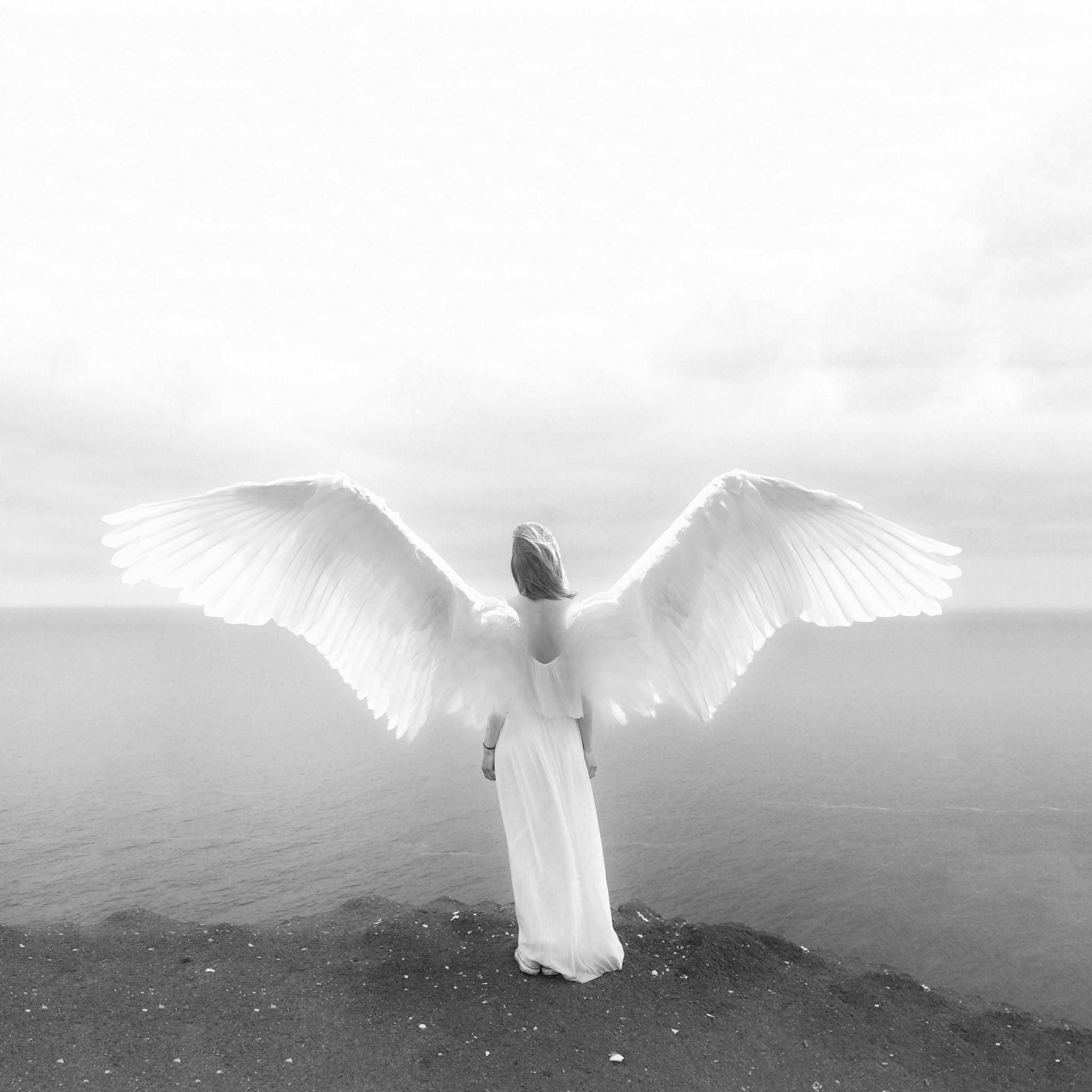 Angel on the Edge B&W, a black-and-white fantasy art photograph depicting a majestic angel with a broad wingspan standing on a cliff, overlooking a serene ocean horizon with a tranquil sky, emphasizing the ethereal, celestial, and spiritual qualities in a monochrome setting.