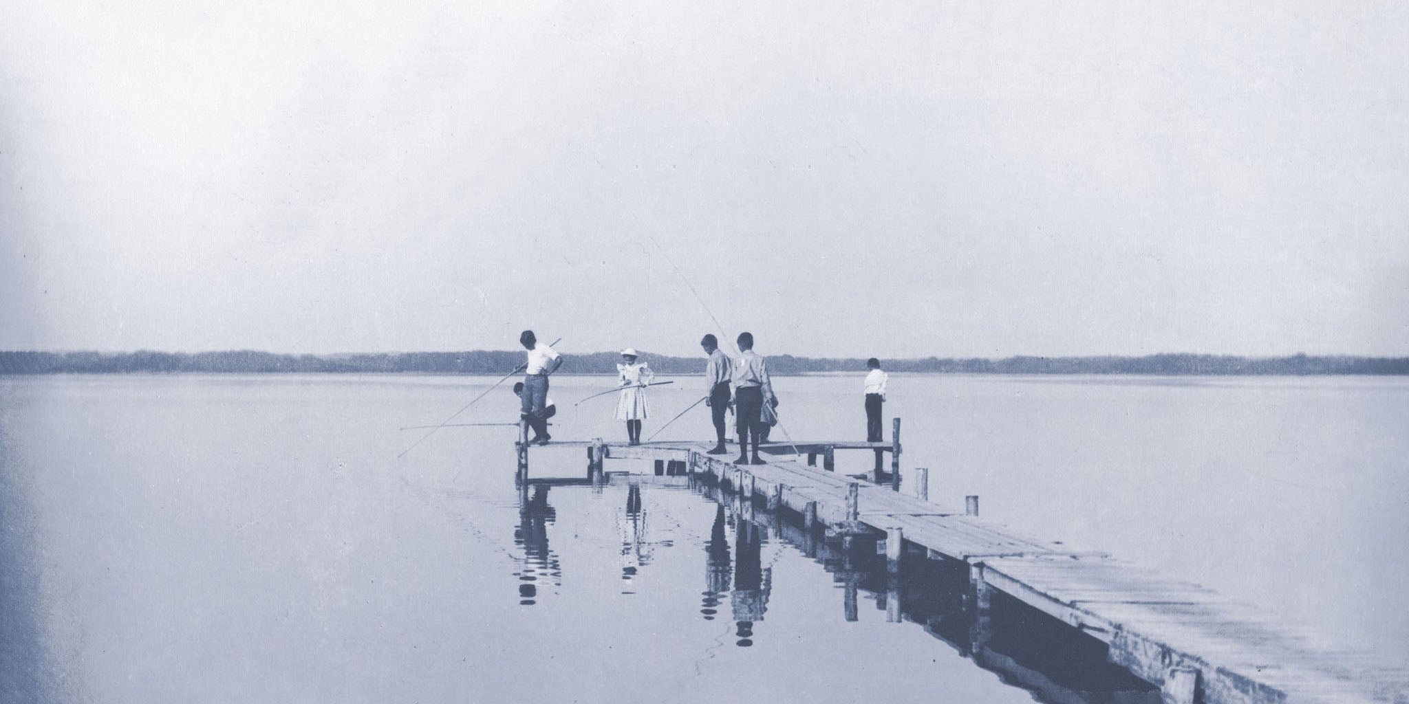 Vintage black and white photograph of children fishing from a wooden dock, capturing a serene moment by the water with reflections and a tranquil sky.